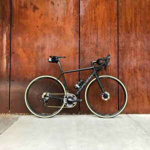 Black road bike with tan tires against a rusted metal wall.