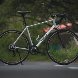 Silver road bike with black handlebars and wheels on a paved road, surrounded by greenery.