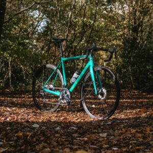 A teal road bike stands on a leaf-covered forest path, surrounded by trees in dappled sunlight.